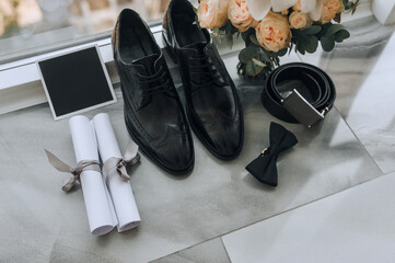 Groom's shiny black shoes, belt, bow tie, scrolls and bouquet stand near window glass against...