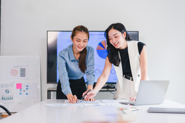 Two businesswomen discussing data analysis, smiling and collaborating at modern office table with...