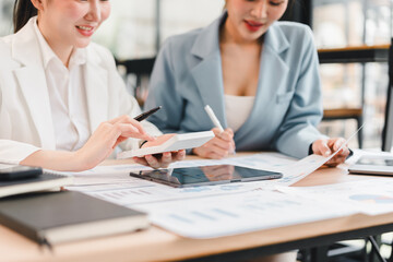 Obraz premium Two businesswomen in formal attire discussing financial documents and digital tablet at modern office desk, collaborating on project with charts and reports, showing teamwork and focus