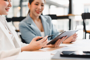 Fototapeta premium Two businesswomen in formal attire discussing work while using digital tablet at modern office, smiling and collaborating on project, with documents and laptop on desk