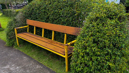 This image features a rustic park bench with a yellow metal frame and wooden slats, placed in front...