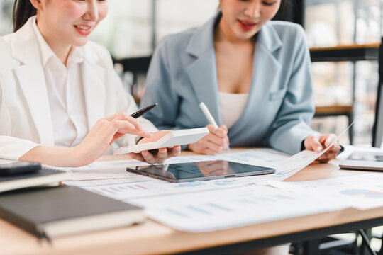 Two businesswomen in formal attire discussing financial documents and digital tablet at modern office desk, collaborating on project with charts and reports, showing teamwork and focus