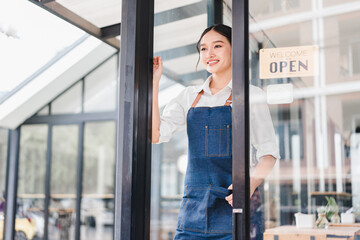 Young Asian woman wearing denim apron standing at glass door of modern cafe, smiling and welcoming customers, sunlight streaming in, cozy atmosphere, small business concept
