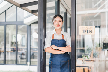 Young Asian woman wearing denim apron standing at entrance of modern cafe, arms crossed, smiling confidently, welcoming customers, glass door with open sign, bright and inviting atmosphere