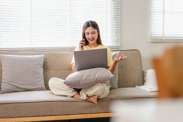 Young woman sitting on sofa with laptop, talking on phone, working from home, casual and relaxed atmosphere, natural light from window, comfortable living room