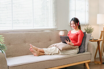 Young woman relaxing on sofa with tablet, wearing red headphones, holding coffee mug, enjoying...