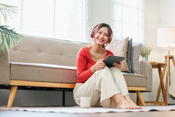 Young woman with headphones sitting on floor, holding tablet, smiling, relaxing in bright living...