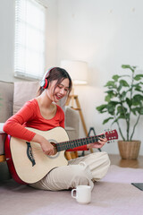 Young woman Asian sitting on floor playing acoustic guitar wearing headphones casual home setting natural light relaxed and joyful mood coffee mug nearby indoor plant decor