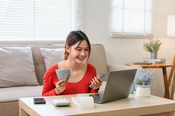 Young woman smiling holding cash and using laptop at home financial success online business remote...