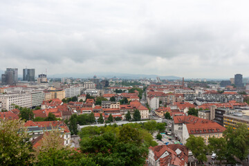 Fototapeta premium An expansive elevated view captures the cityscape of Ljubljana, on a cloudy day. Red-tiled rooftops dominate the foreground, showcasing the charming historic architecture of the old town