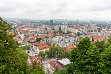 Obraz premium An expansive elevated view captures the cityscape of Ljubljana, on a cloudy day. Red-tiled rooftops dominate the foreground, showcasing the charming historic architecture of the old town