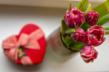 Top photo of bouquet of vibrant pink tulips in focus with a blurred red heart-shaped gift box in a...