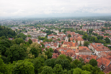 Obraz premium An expansive elevated view captures the cityscape of Ljubljana, on a cloudy day. Red-tiled rooftops dominate the foreground, showcasing the charming historic architecture of the old town