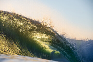 A beautifully vibrant backlit emerald gold coloured glassy surfing wave breaking over a shallow reef. Captured from the ocean's surface on sunrise.