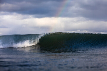 A wave curls and breaks along a sandy beach under a cloudy sky with a faint rainbow in the...