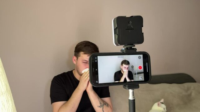 A man records a video on his phone using a tripod at home, but suddenly experiences an allergic rhinitis attack. He starts sneezing and sniffling during filming.