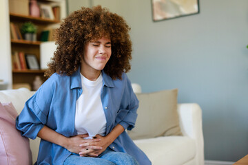 Curly-Haired Woman with Pained Expression Sitting in Living Room Holding Stomach