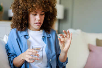 Young Woman With Afro Hair Holding Glass Of Water And Medication Wearing Blue Denim Shirt On Sofa