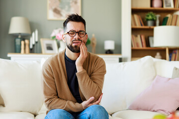 Young Man in Casual Outfit Sitting on Sofa Holding Jaw in Discomfort in Well-Lit Living Room