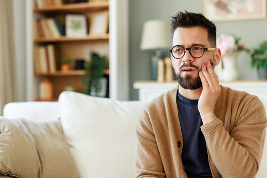 Young Man in Beige Cardigan Holding Jaw in Bright Living Room While Experiencing Pain