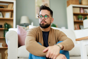 Thoughtful Young Caucasian Man Sitting in Cozy Living Room Wearing Glasses and Casual Clothing