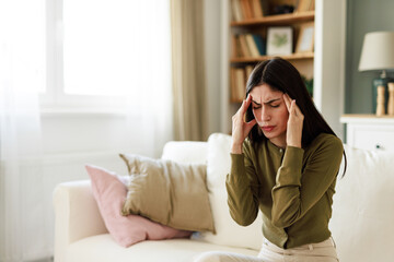 Young Woman in Green Sweater Sitting in Bright Living Room Holding Head in Pain