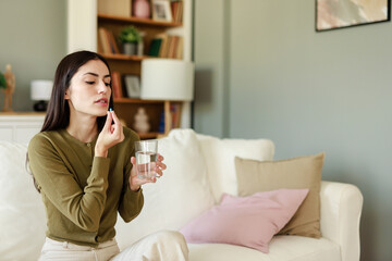 Young Woman With Long Dark Hair Taking Medicine With Water Glass While Sitting On White Couch At Home