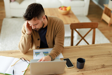 Frustrated Caucasian Man In Beige Sweater Reviewing Financial Documents At Home Office With Coffee And Laptop
