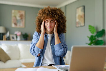 Young Woman with Curly Hair Holding Head in Pain While Working from Home in a Comfortable Living Room