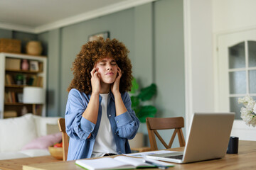 Young Professional Woman With Curly Hair Holding Head In Pain During Remote Work At Bright Home Office