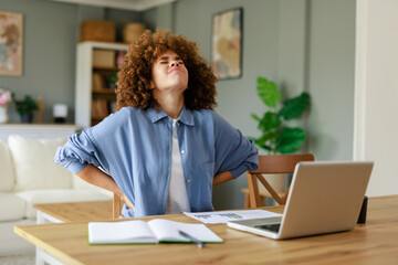 Young Woman with Curly Hair Experiencing Back Pain While Working from Home Indoors in a Brightly Lit Room