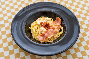 spaghetti carbonara with grilled and crispy bacon ham for noodles cuisine and Italian food in black plate or dish on yellow white brown table or tablecloth by top view at food court or restaurant