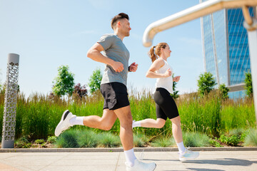 Couple jogging together in a sunny urban park, surrounded by greenery and modern architecture...
