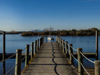 Obraz premium Straight wooden pier with rope railings points to a small white motorboat moored on calm water. Low brush lines the far bank and distant hills sit under a clear blue sky, giving a quiet riverside mood