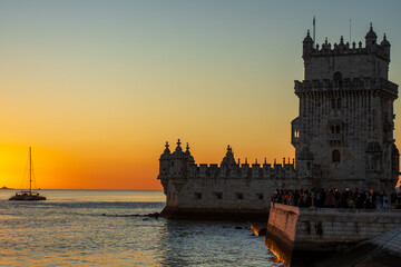 Torre de Belém stands against an orange sky as the sun drops over the Tagus. A catamaran sails...