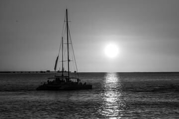 Black-and-white photo of a catamaran sailing across the Tagus at sunset. The boat and its mast form a dark outline against the glowing horizon, with the Vasco da Gama Bridge faint in the distance. 