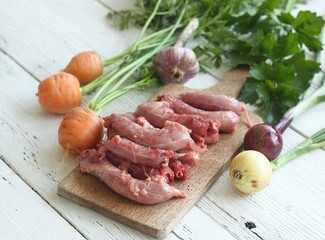 Raw chicken necks on a cutting board. Fresh baby carrots, onions and garlic on a white wooden table.