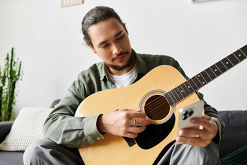 Young man enjoying music at home while playing guitar and checking his phone for lyrics
