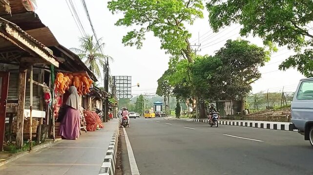 Suasana jalan di perkotaan yang begitu lengang, dengan mobil dan sepeda motor yang lalu lalang dan terlihat ada yang jualan di pinggir jalan. 