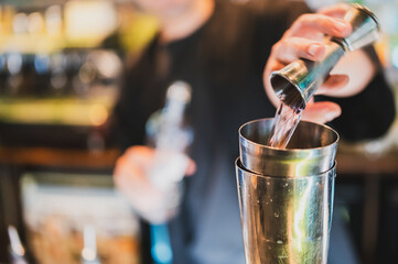 Bartender pouring liquid from jigger into cocktail shaker in bar setting. Close-up of hands and tools, showcasing precision in professional drink preparation.