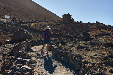 Woman climbing a rugged volcanic trail toward the summit of Pico del Teide surrounded by dark lava...