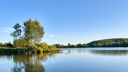 Tranquil summer landscape with tree reflections in a clear lake, forested shoreline and blue sky. Perfect for nature, relaxation, and eco-themed concepts.