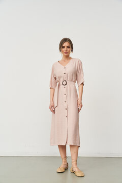 Woman in a light pink belted dress with button details stands confidently, wearing beige shoes. She has brown hair styled in an updo and delicate makeup. The style is elegant and simple