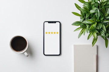 A smartphone displaying a five-star rating sits on a white desk next to a cup of coffee, a green plant, and a closed notebook.