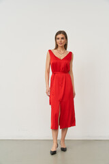 Woman in a sleeveless red dress with a v-neck and belt, wearing a necklace. She's standing in a...