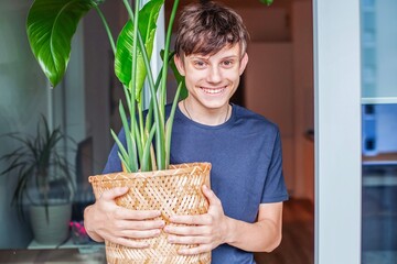 Cute cheerful teenager bought and brought home a green houseplant ,young man holding potted plant