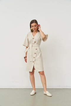 Elegant woman in a light buttoned dress stands with a relaxed pose, hand touching ear, wearing light loafers. Her other hand rests on her hip. White backdrop enhances minimalistic style