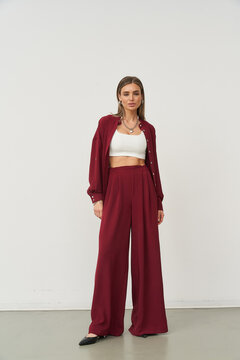 Woman stands confidently in chic outfit, featuring burgundy blazer and pants paired with a white crop top. Minimalist jewelry and straight hairstyle complete the stylish look