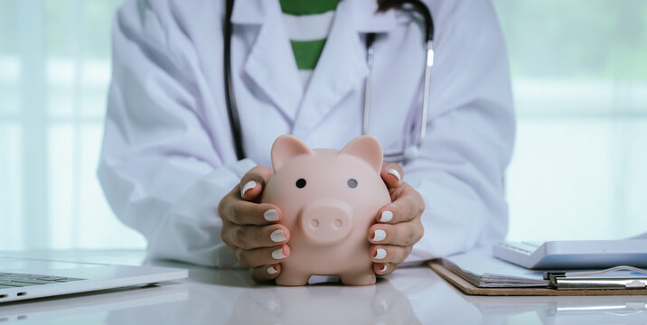 Close up young female general practitioner putting coin in piggy bank, managing hospital budget using laptop accounting applications, medical insurance finances, healthcare money savings concept.