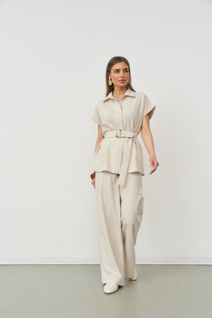 A woman in a beige outfit poses confidently, wearing a short-sleeved top with a belt and wide-legged trousers. She stands against a plain backdrop, wearing statement earrings
